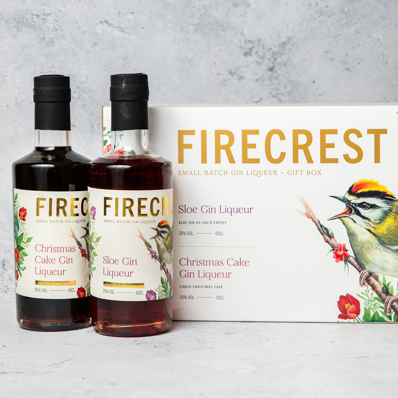 Two bottles of Firecrest Sloe Gin and Christmas Cake Gin Liqueur with a box in the background on a gray surface.